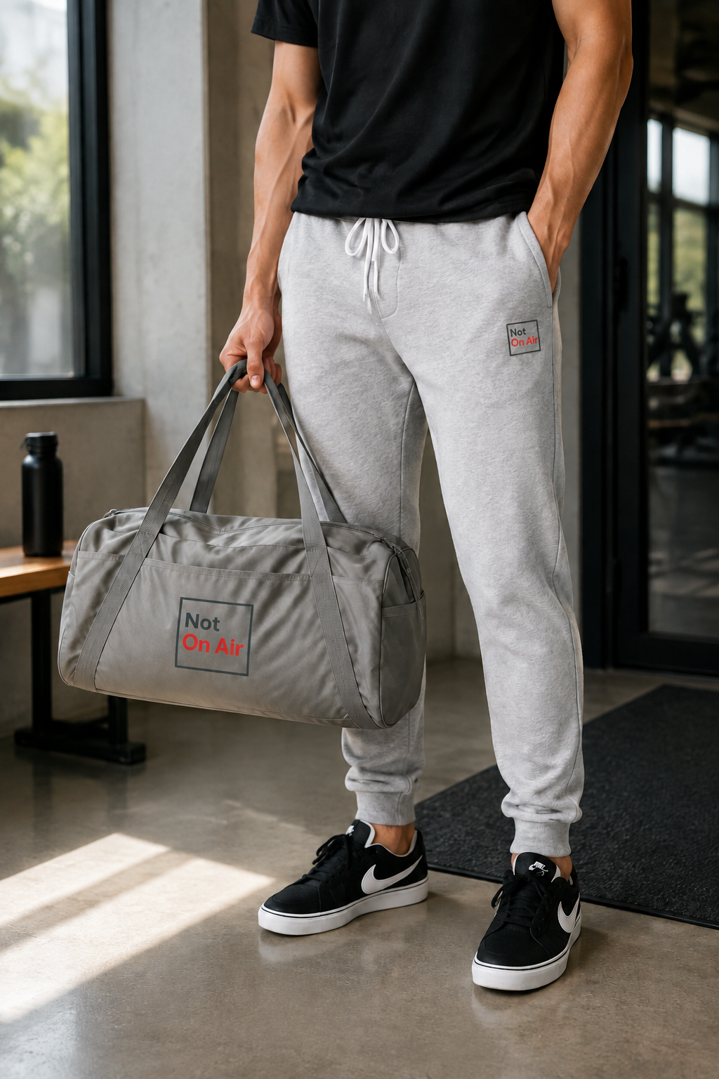 Cropped model in a black shirt and gray Not On Air embroidered joggers carrying a matching gray duffel.