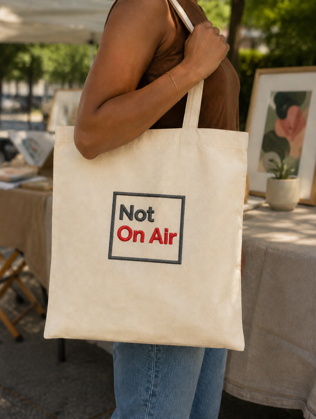Faceless model carrying a natural canvas Not On Air tote bag with realistic gray and red embroidery.